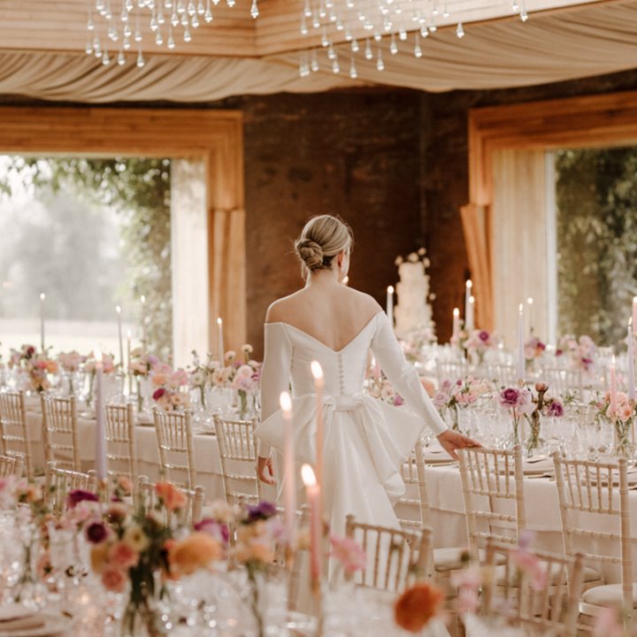 A modern bride in stunning scoop back wedding dress with huge bow surveys her gorgeous romantic soft pink wedding reception with pretty flowers and candles laid out on glowing tables in the twinkling light of The Gillyflower at a modern wedding at Cotswolds venue Elmore Court