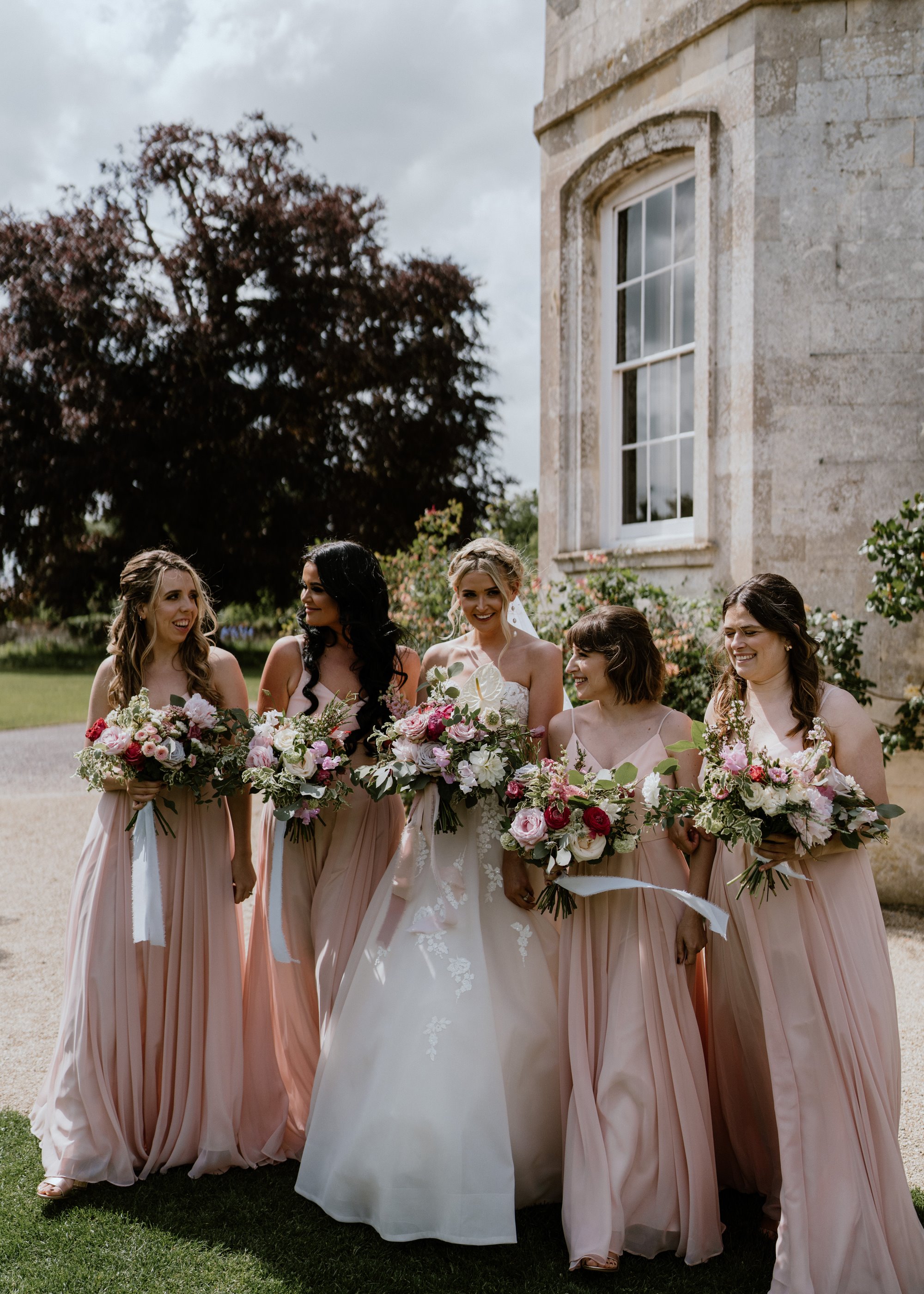 Bridesmaids and bride in romantic pink dresses holding bouquets with ribbons in front of beautiful mansion house wedding venue