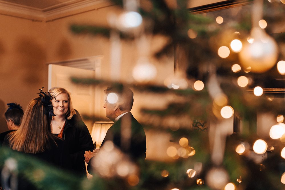 wedding guests seen through the branches of a christmas tree at winter wedding reception in a stately home