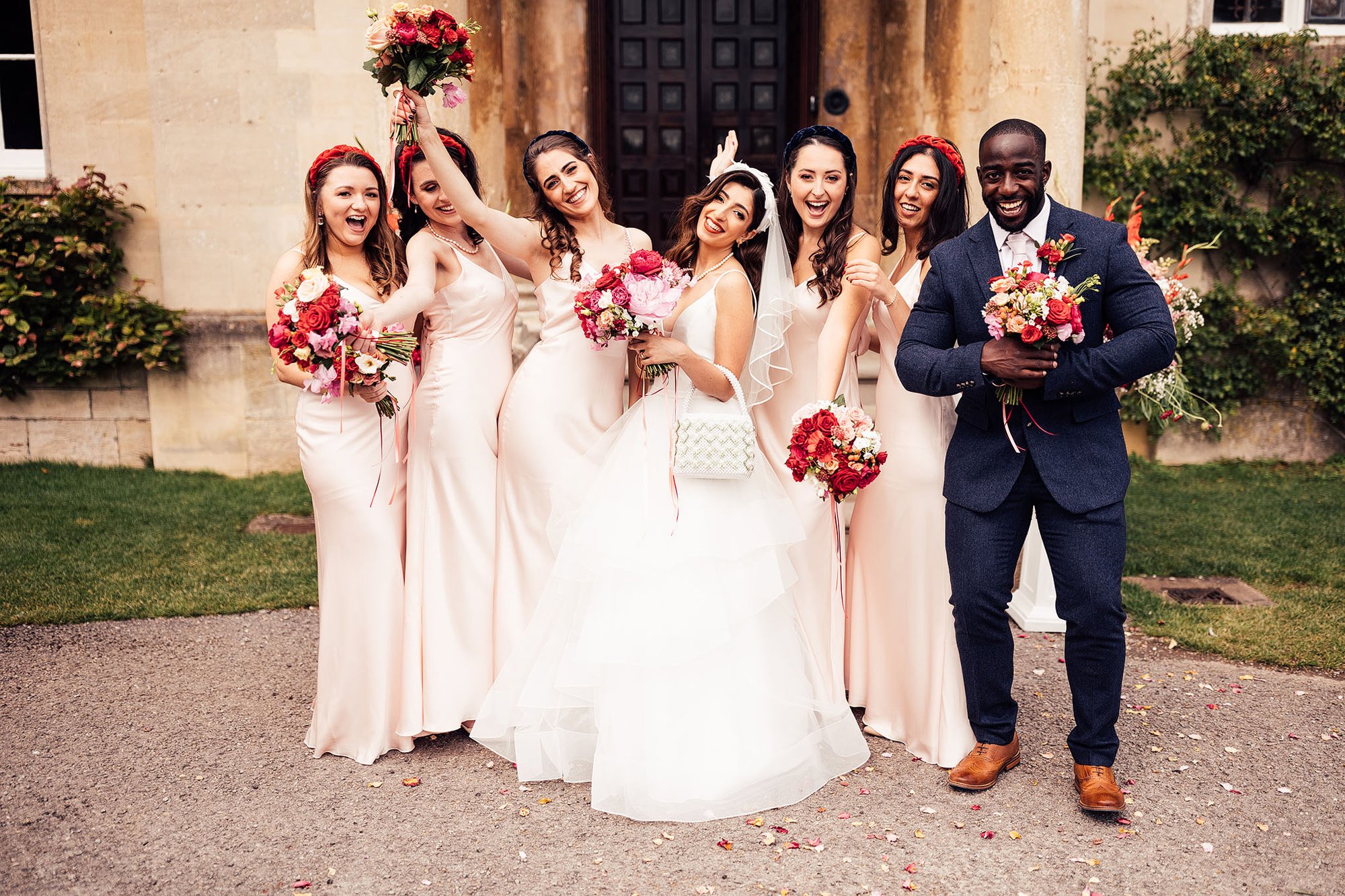 A bridal party with bouquets of red flowers posing with the groom outside a stately home in Elmore