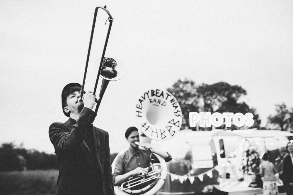 A black and white photo of a brass band playing outside at a wedding in front of a photo booth with sign saying photos in capital letters in the background