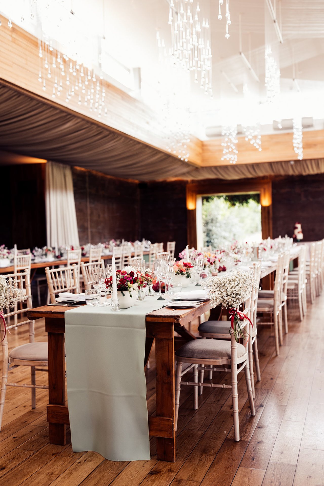 wedding reception with long banqueting tables with pale green linen and pink and red floral details and babys breath decor on chairs
