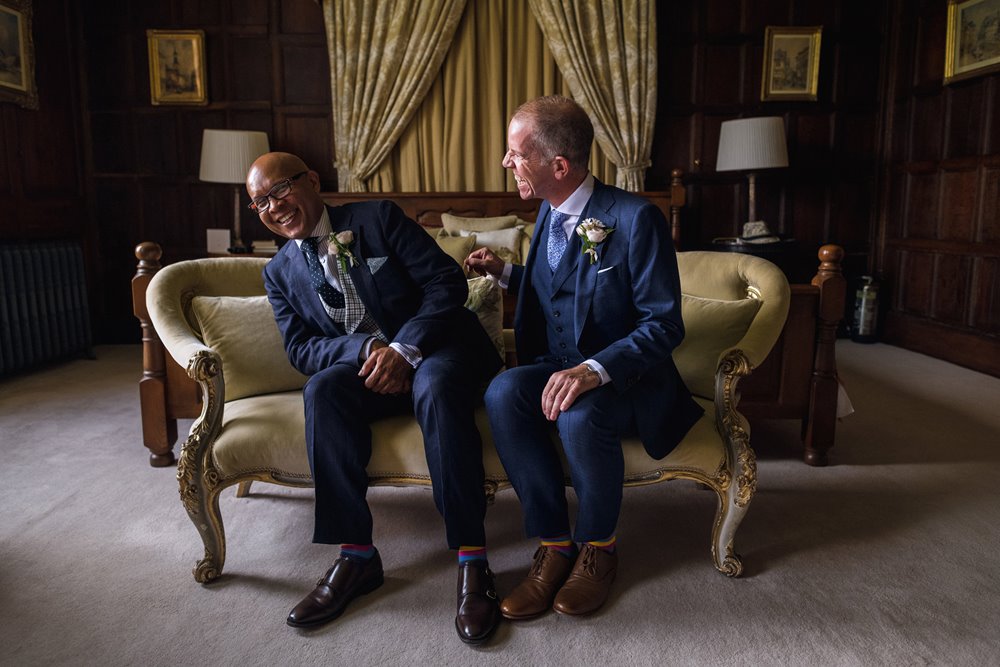 Two grooms laugh sitting on chaise longue of stately home bridal suite elmore court
