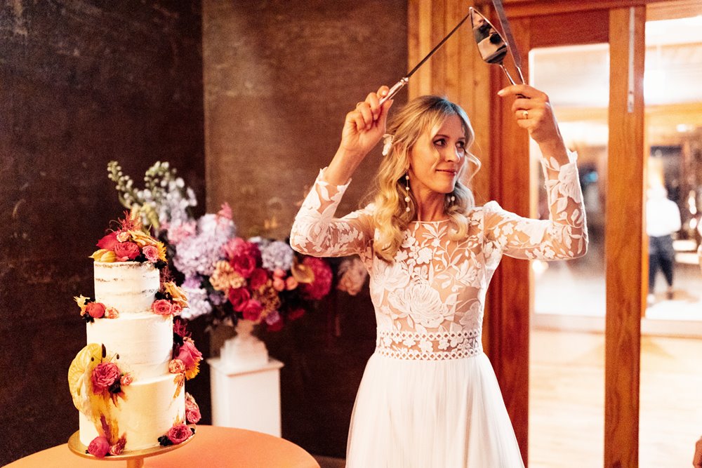 boho bride cuts cake next to colourful wedding flowers and candles at wedding venue in gloucestershire