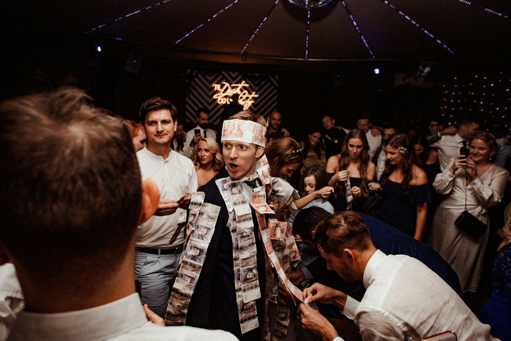 Groom wearing money crown gets money pinned to his jacket by guests during Greek wedding money dance at Elmore Court