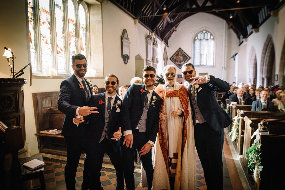 Groomsmen in sunglasses pose with the Vicar of St John the Baptist church at a relaxed church wedding in Elmore