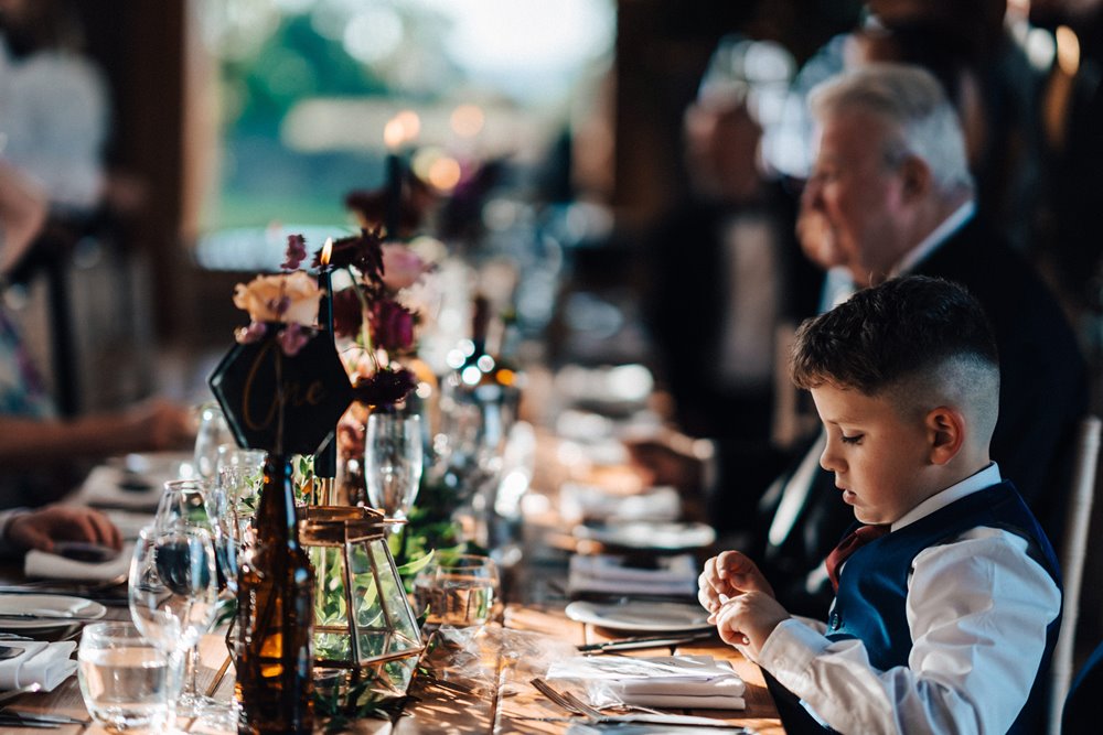 Wedding guests sitting at long banquet tables decorated with black flowers and mismatched glasswear for a dark and moody autumn wedding in the cotswolds