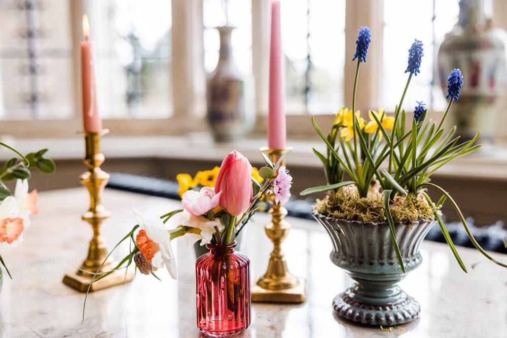 Pretty little spring flowers in tiny vases and bottles and pink candles decorate the piano in stained glass mullion window of wedding ceremony hall at elmore court