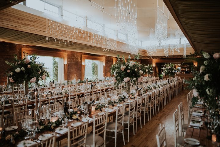 A festival wedding banquet in The Gillyflower at Elmore Court, long banqueting tables are laid with candles, tall flower arrangements and a beautiful twinkling light showers hangs from the ceiling