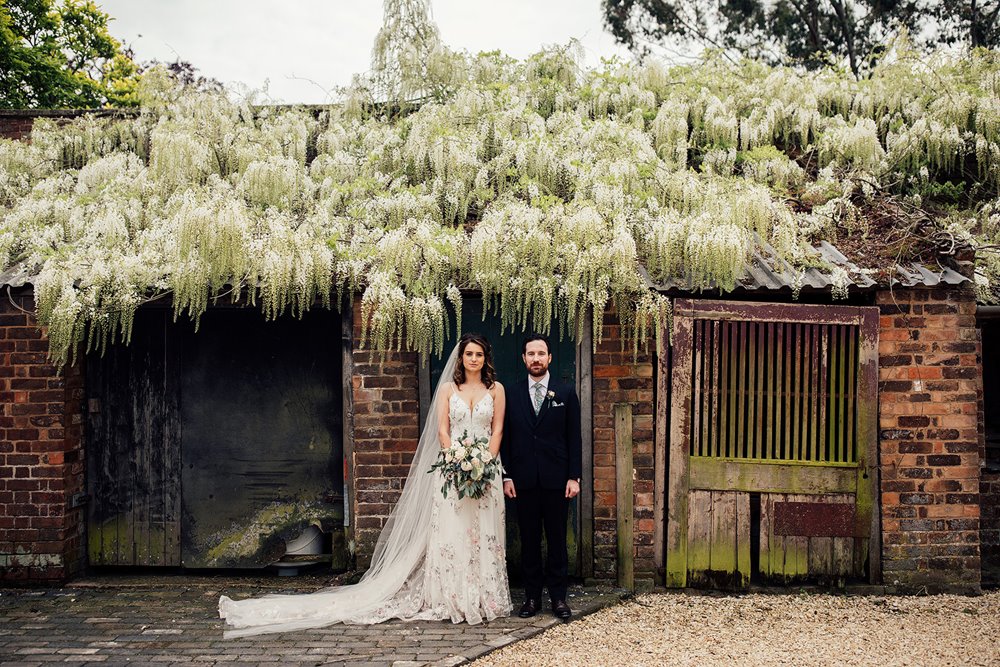 Bride and groom pose in front of beautiful white trailing blossom on a out building at country wedding venue elmore court