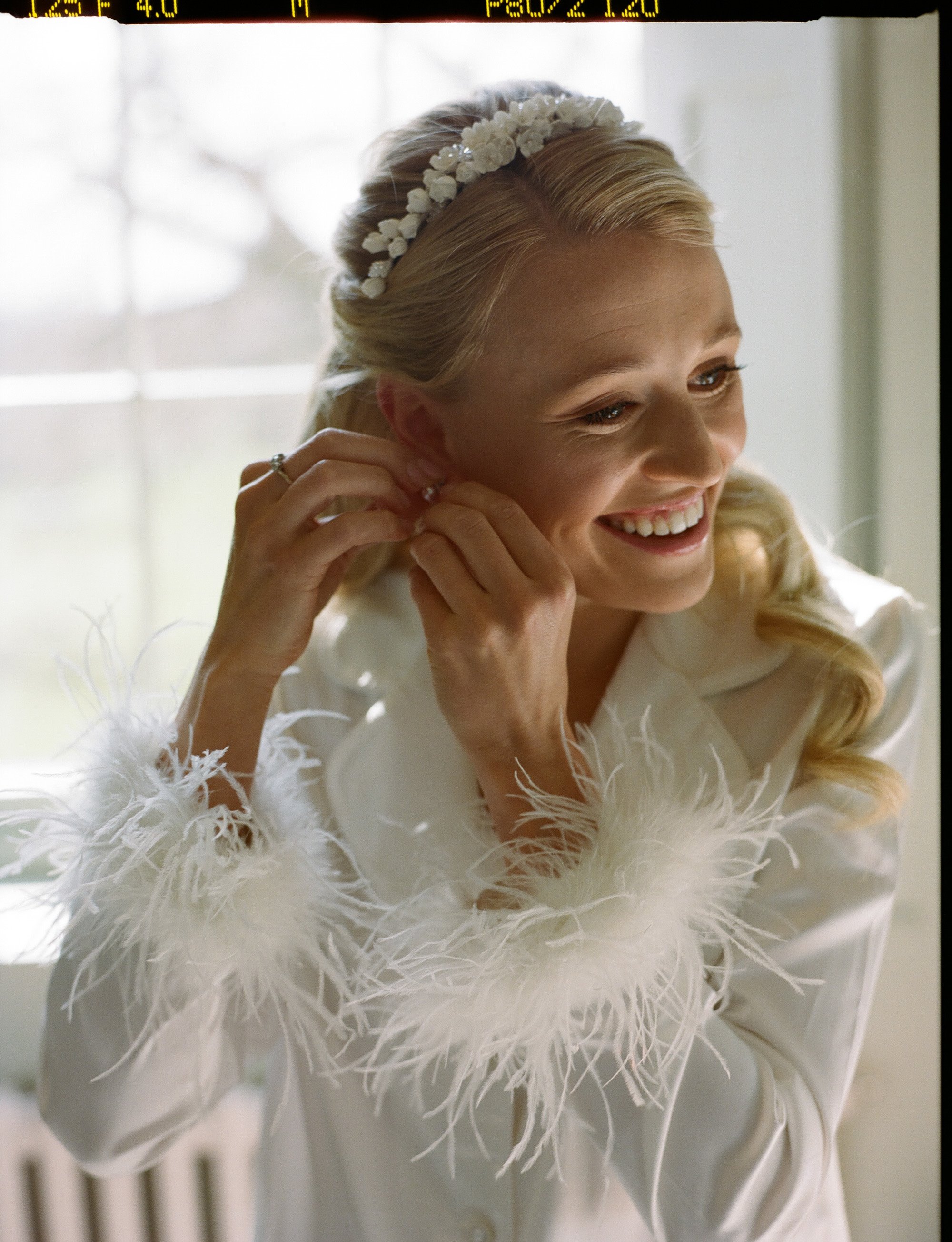 Elegant blonde bride in white feather nightwear and pearl tiara puts on her pearl earrings on her wedding morning at glamorous wedding venue Elmore Court