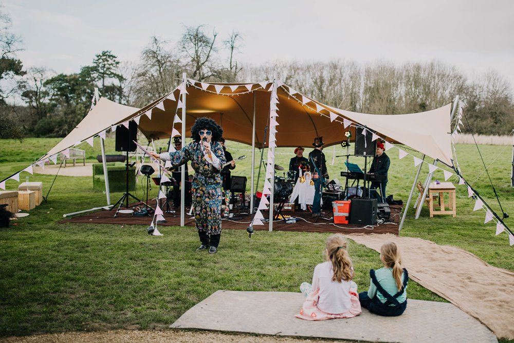 Children watch band play under stretch tent with lead singer dressed up in afro and kaftan for wild wedding fair at elmore court