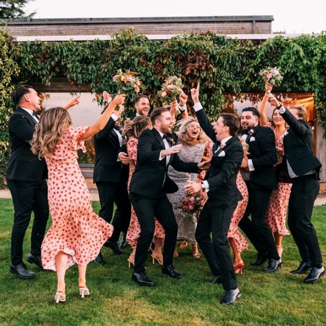 cool Wedding guests and bride and groom jump around in celebration wearing heart dresses in front of sustainable wedding venue covered in greenery