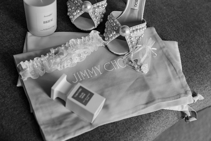 Pearl bridal shoes by jimmy choo and pearl wedding garter laid out with perfume and candle and pearl accessories on wedding day at luxe wedding venue elmore court