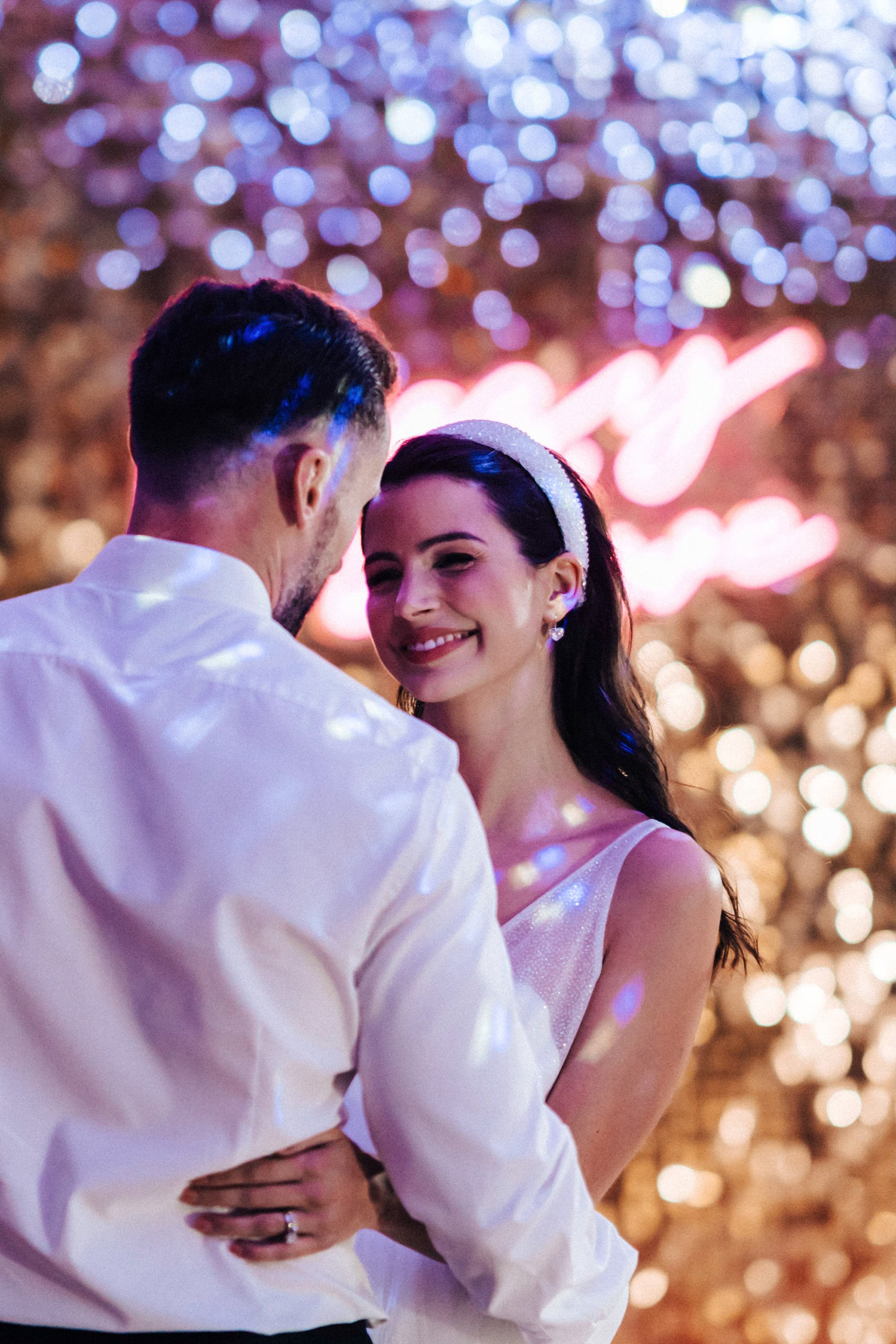 Beautiful bride in pearl earrings and headpiece smiles dances with groom against sequin backdrop