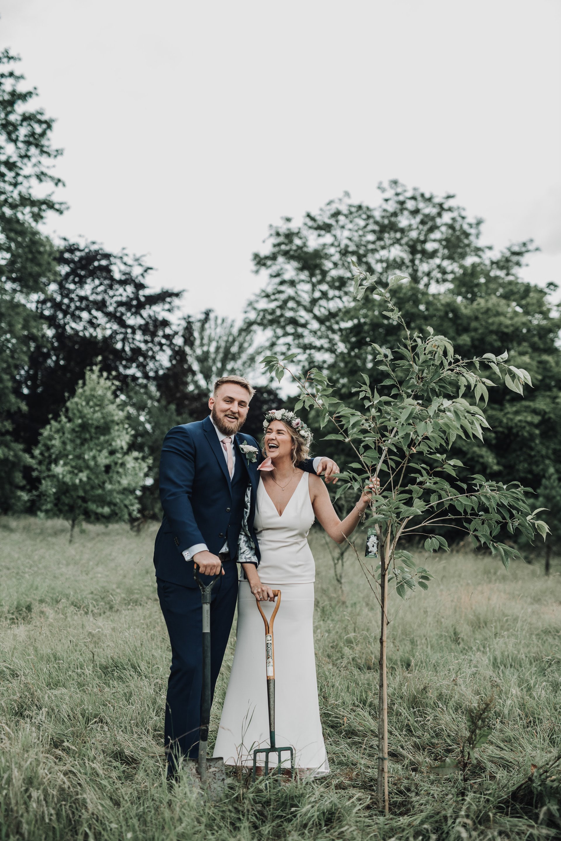 Bride and groom stand with the tree they just planted as part of their micro wedding ceremony during corona pandemic at sustainable wedding venue elmore court in cotswolds