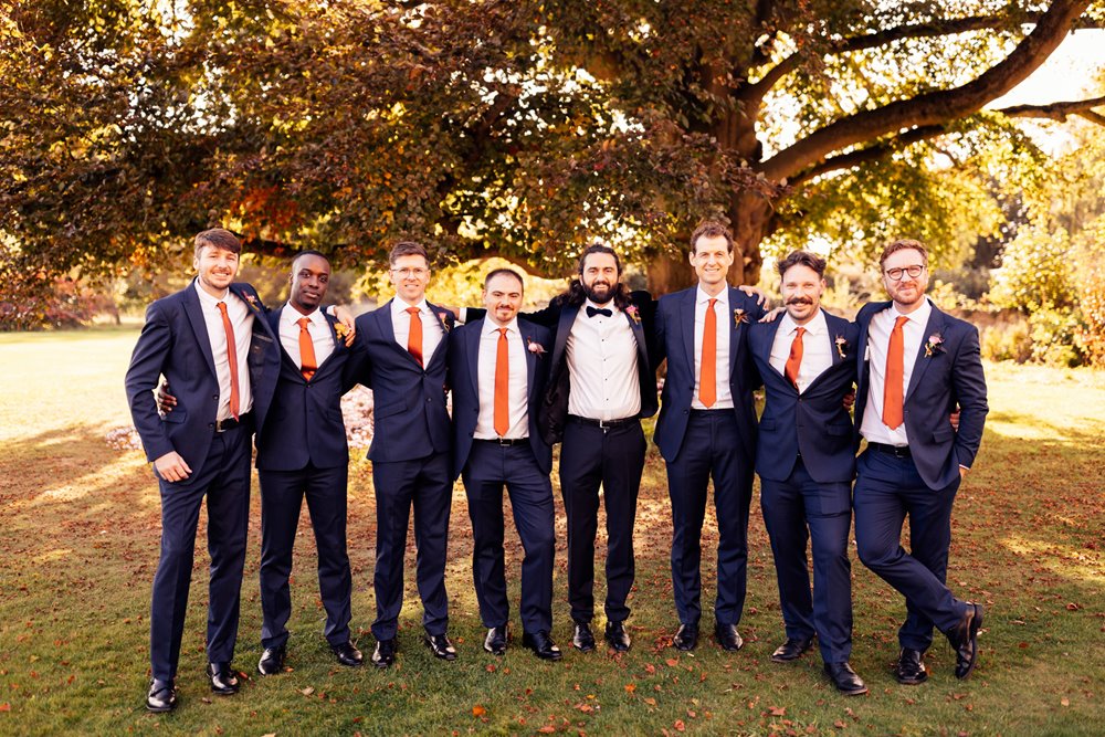 Groomsmen in suits with bright orange ties under beech tree outside stately home