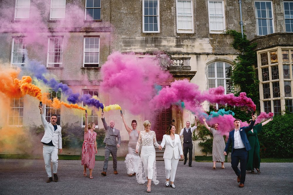Lesbian wedding party surrounded by clouds of colourful smoke in front of their stately home wedding venue