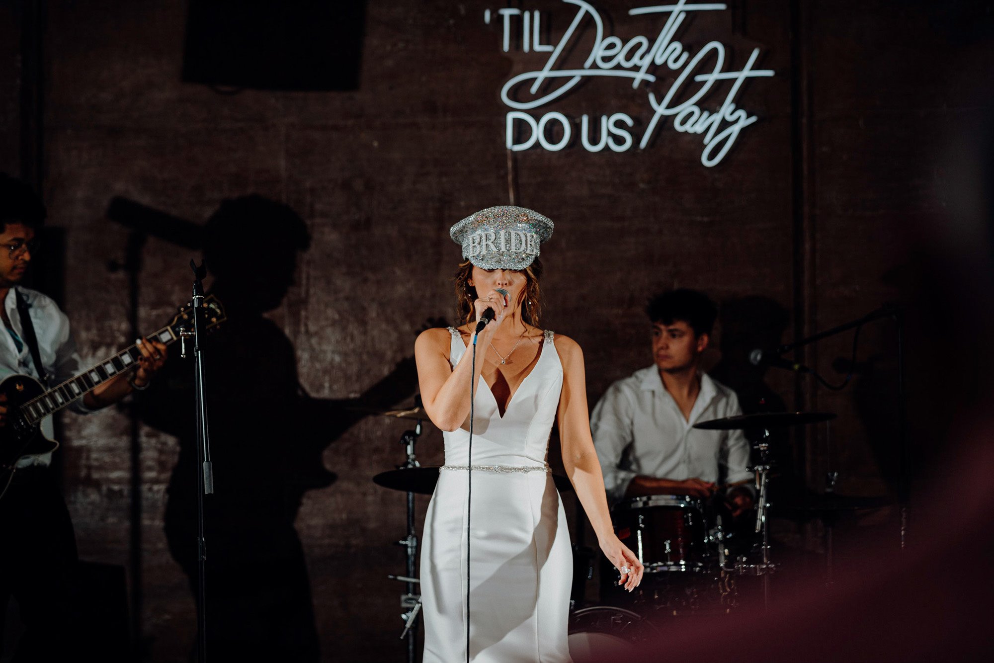 Rockstar bride on stage wearing sparkly hat with bride in pearls and death do us party sign at party wedding venue elmore court