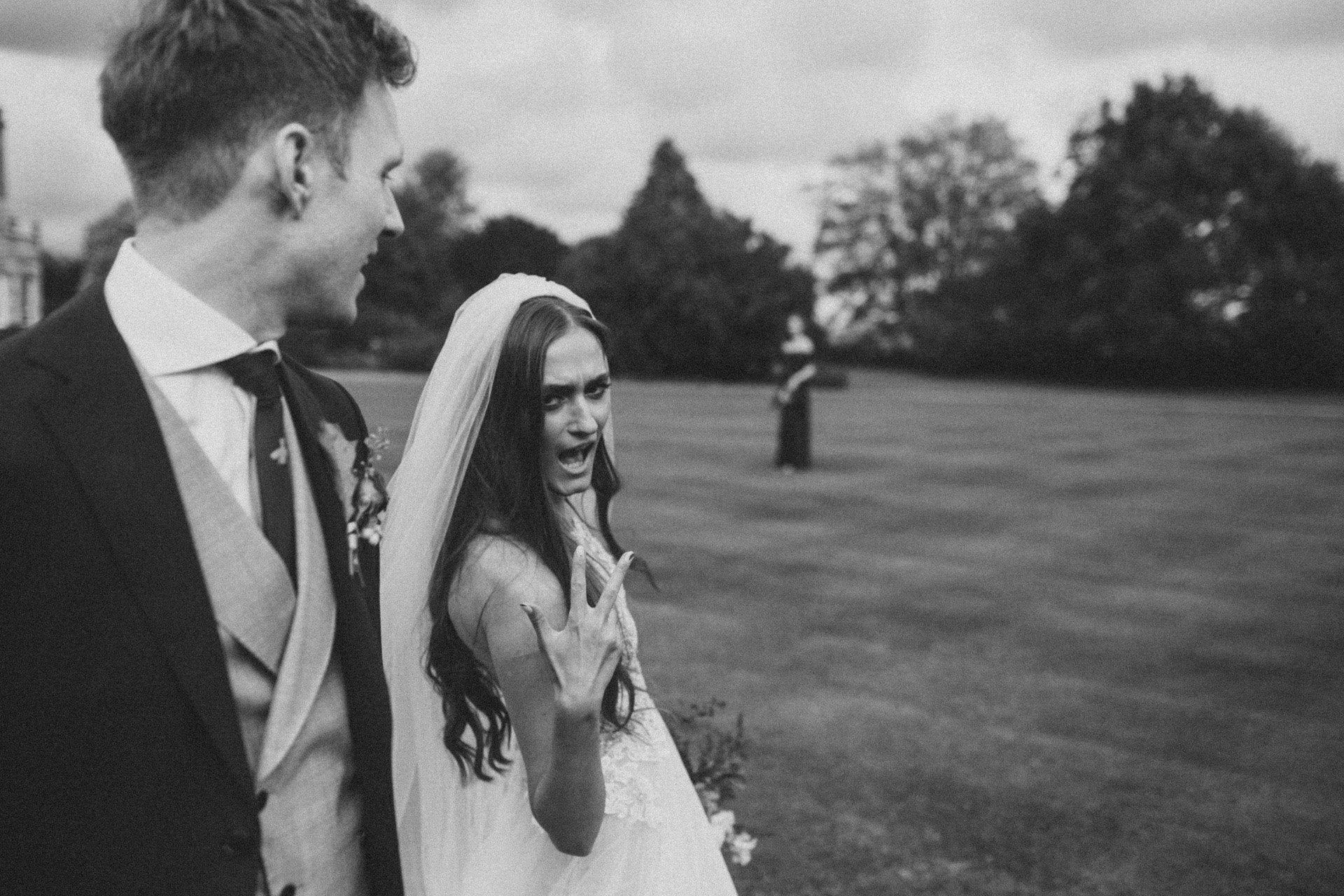 Cool modern boho bride pulls v sign at camera walking next to her groom on the lawn of her mansion wedding venue