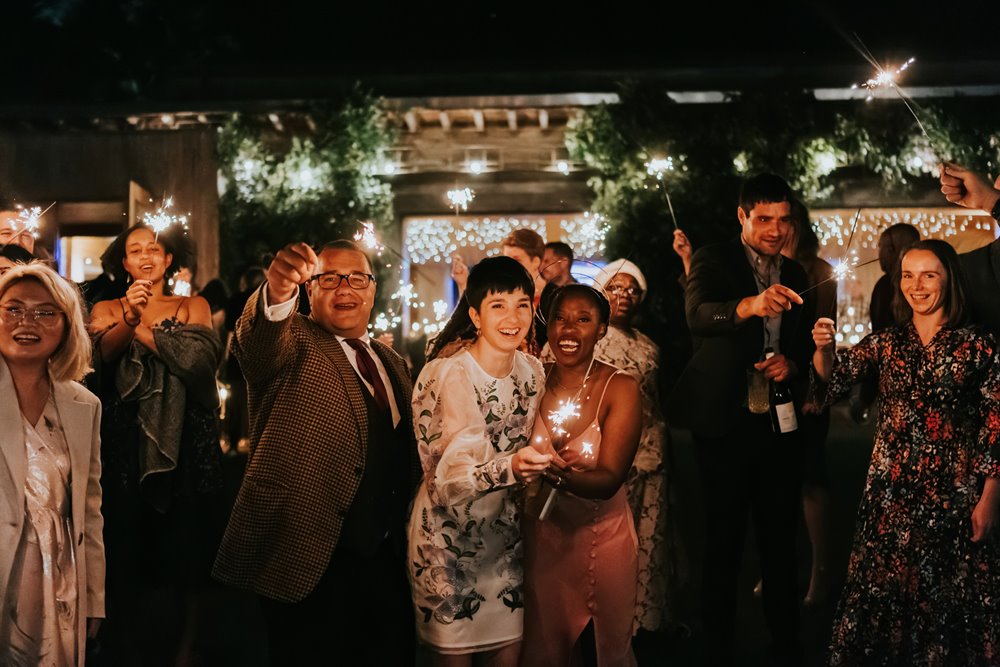Two brides surrounded by their friends holding sparklers on their magical multicultural same-sex wedding day at elmore court in UK