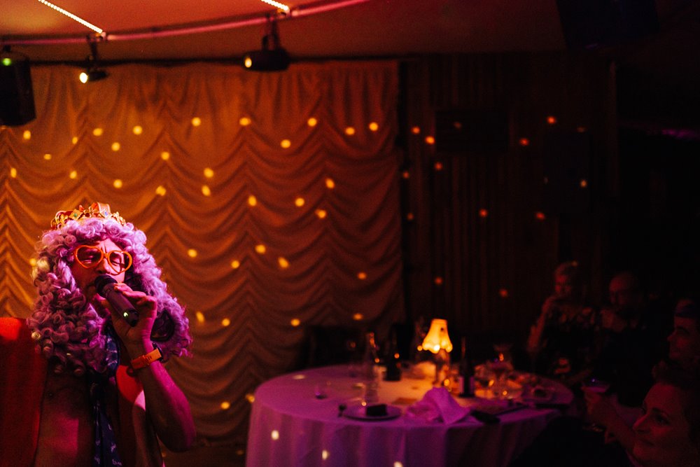 Live music cabaret show. Front man sings into a microphone gets down from stage wearing curly wig and glasses