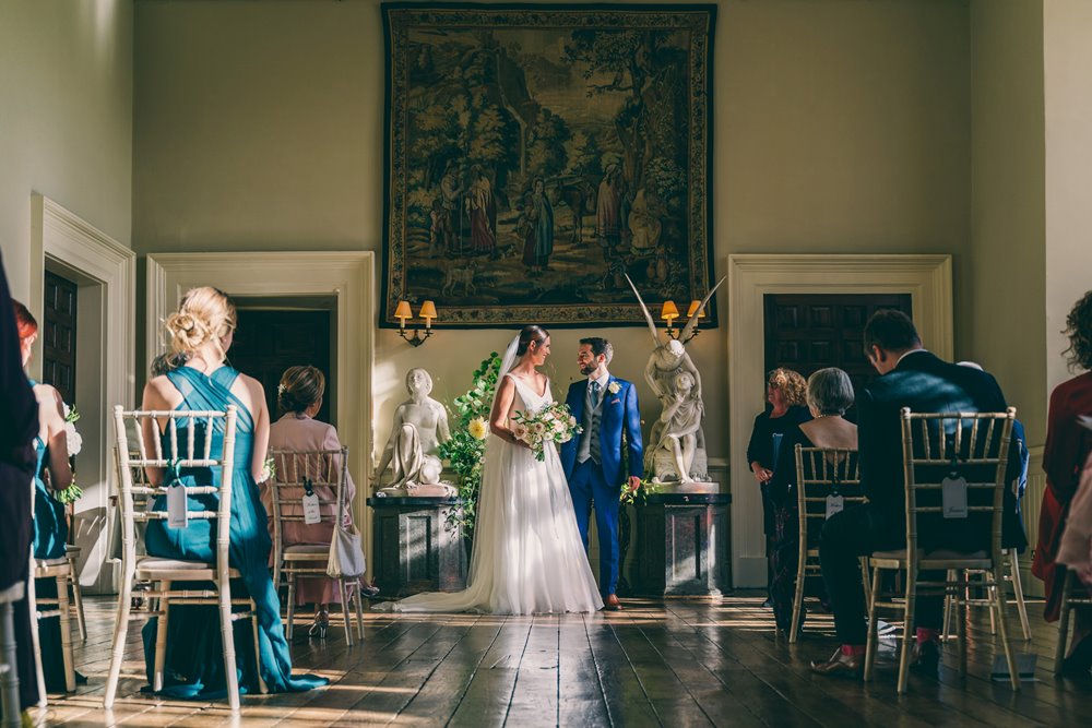 Micro wedding ceremony in stately home hall. Bride and groom stand at end of aisle with a small number of guests watching