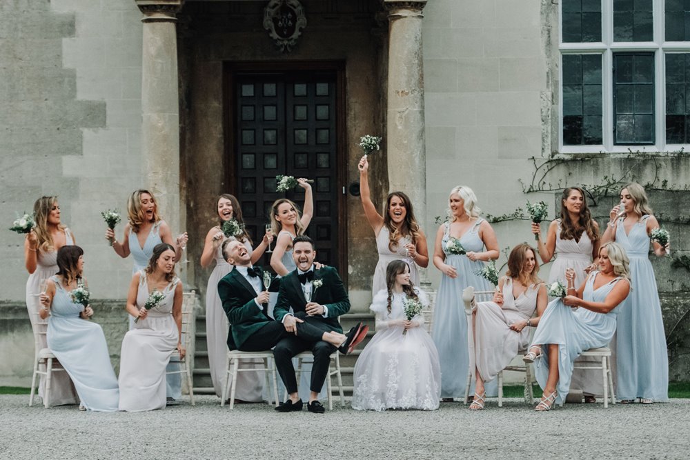 Gay grooms pose and laugh with their 13 bridesmaids of mega wedding party at mansion house elmore court at this luxe same sex celebration