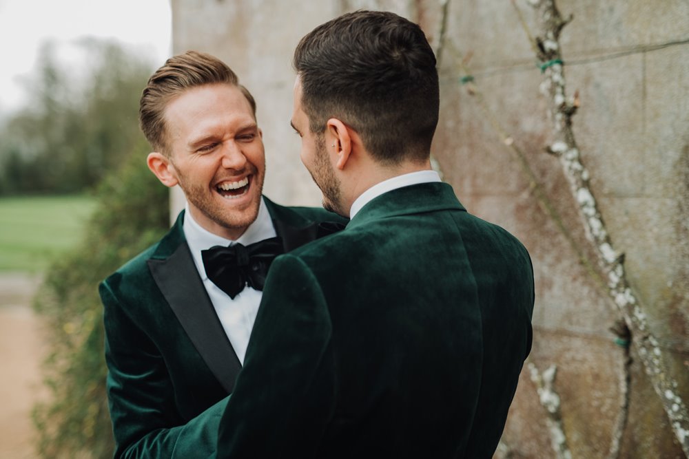 Two grooms wearing green velvet tuxedos grin at each other outside their mansion house wedding venue on their perfect luxe gay wedding day