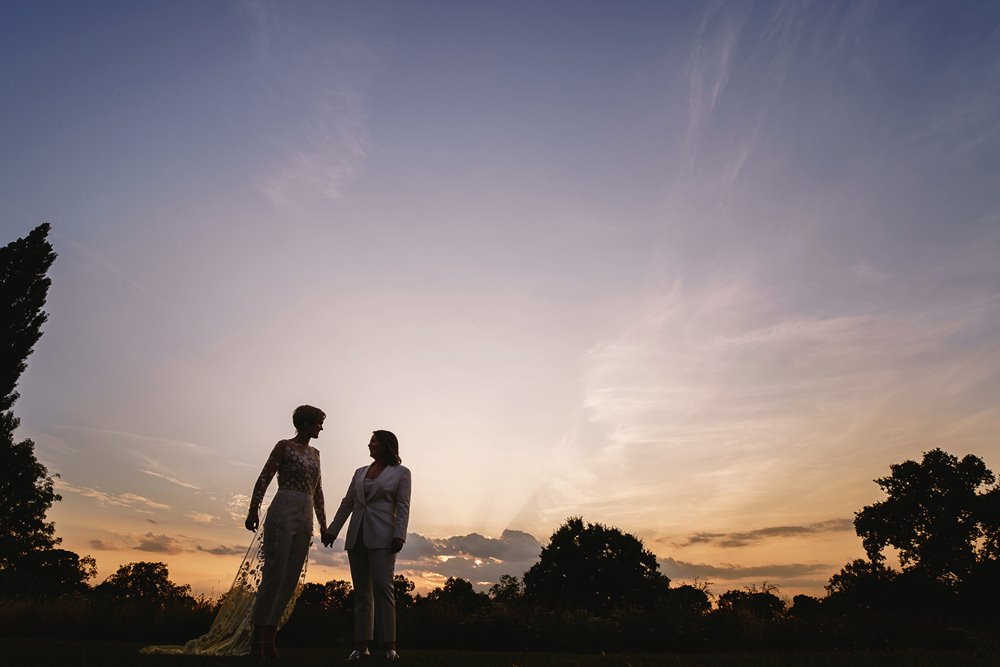 Lesbian brides hold hands and walk into the sunset on their beautiful wedding day at elmore court