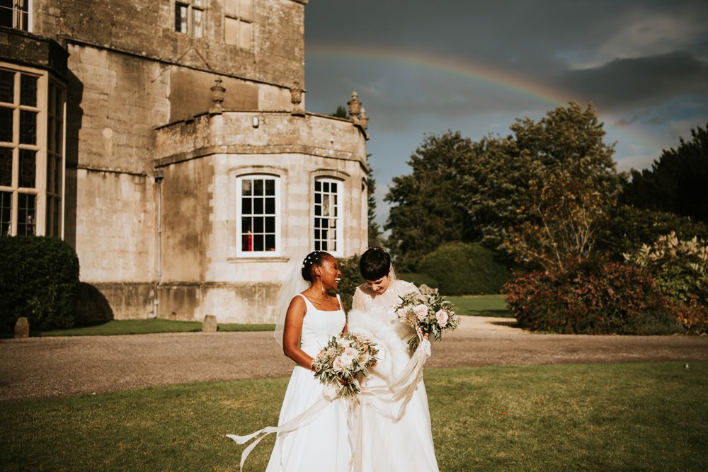 Two brides in white wedding dresses stand outside their stately home wedding venue underneath a rainbow blessing their same sex marriage in Cotswolds countryside