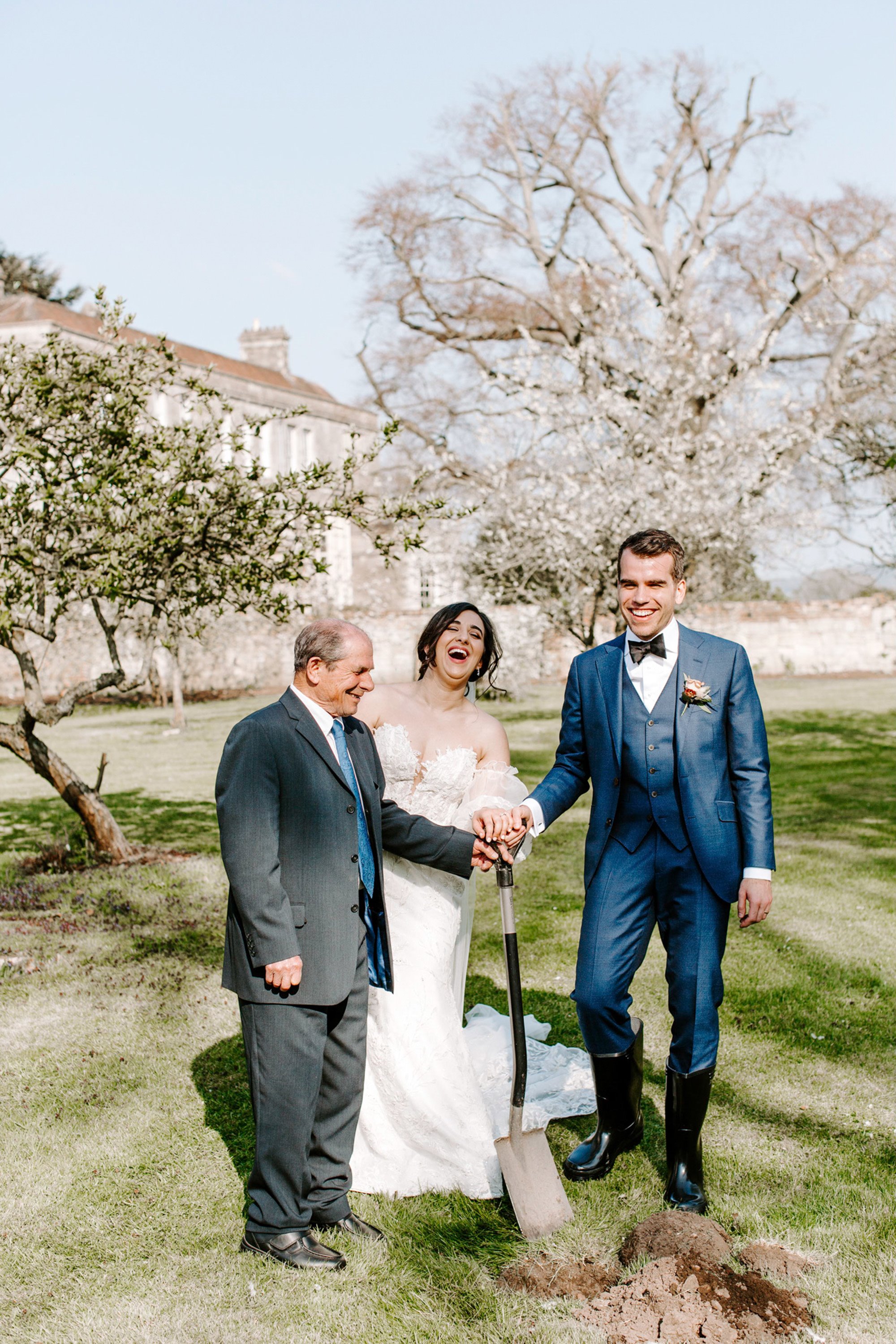 Bride, groom and father of bride all hold handle of spade to dig the first earth as part of their tree planting ceremony outside wild wedding venue elmore court in the cotswolds