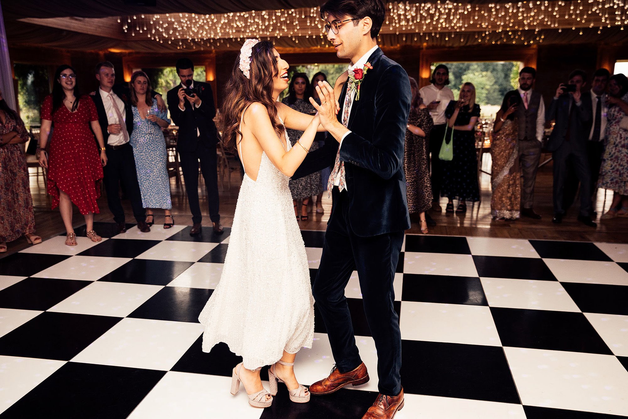 Modern bride wearing pearl reception dress and groom in blue velvet suit dance on chequered dance floor with twinkling lights above