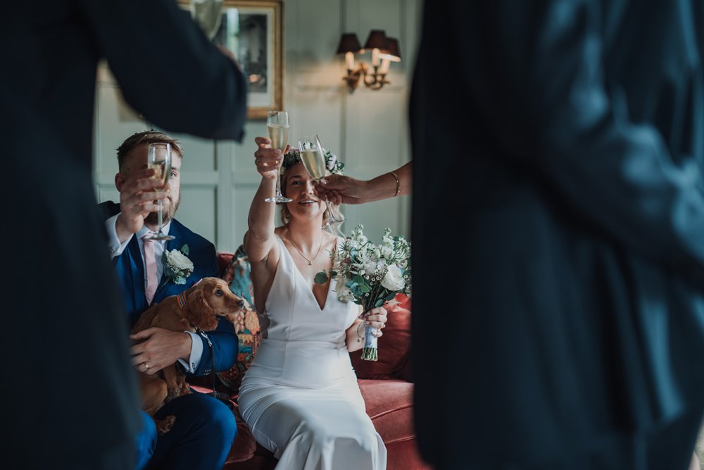Micro wedding couple with dog bridesmaid cheers champagne glasses in wood panelled drawing room at elmore court