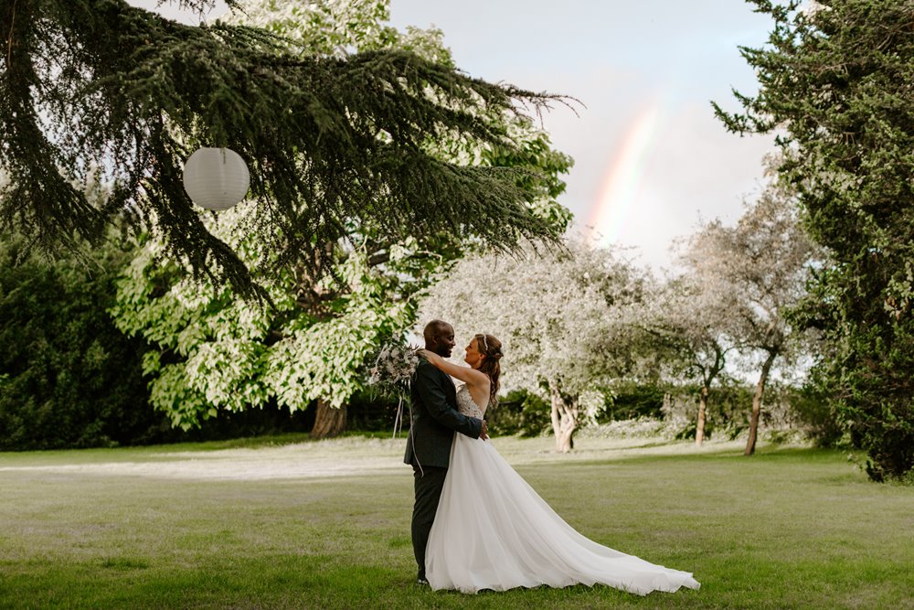 Couple embrace under the cedar tree with a rainbow appearing in the sky at a magical outdoor wedding