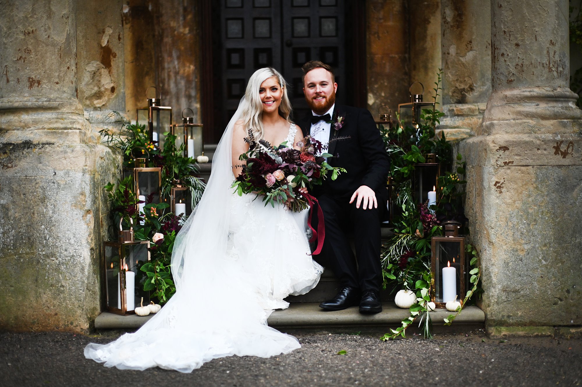 Bride and groom sit on the steps of their unusual mansion house wedding venue surrounded by rustic autumnal decor for their halloween wedding