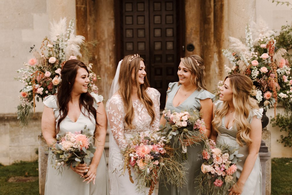 Modern boho luxe bride and bridesmaids stand outside country house wedding venue holding soft pink bouquets with trailing greenery