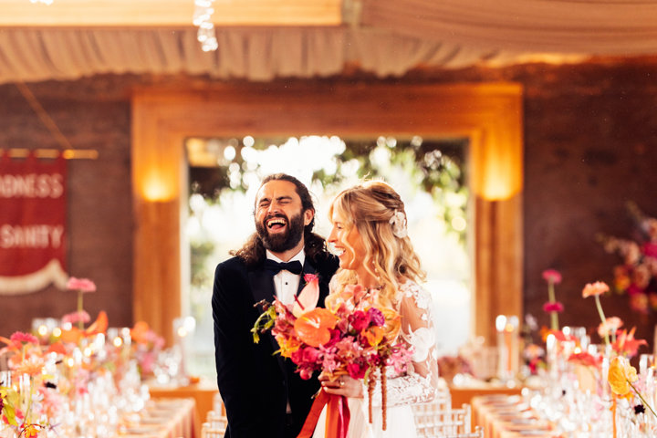 Fun loving boho bride and groom laugh with joy at their colourful wedding reception with bright flowers in the cotswolds