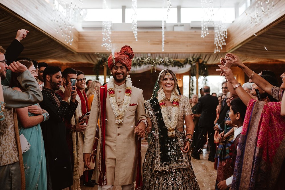 Bride and groom leave ceremony room with mandap behind surrounded by guests at their indian wedding venue