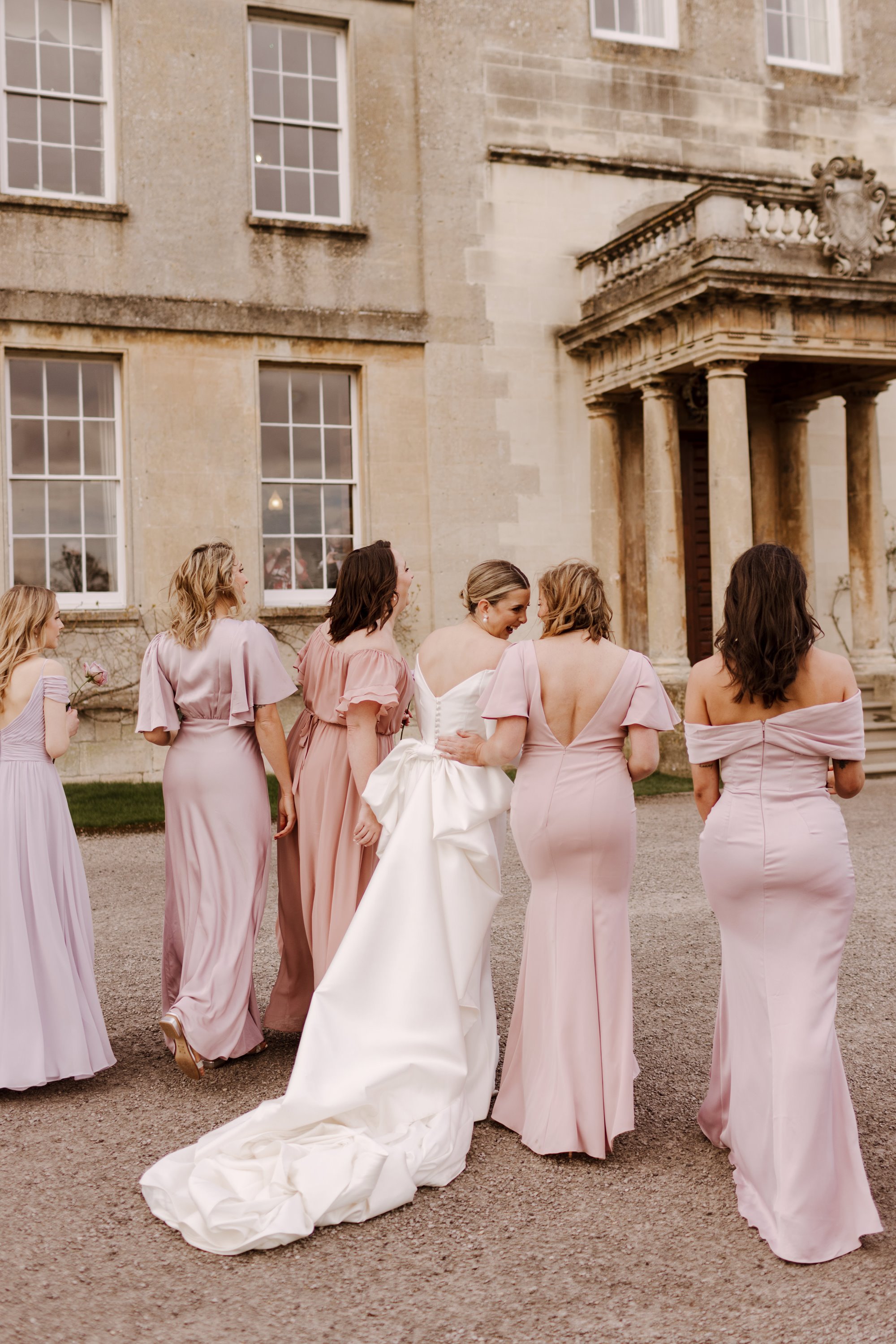 Bride in dress with huge bow and bridesmaids in pink dresses figure hugging with backs to the camera in front of stately home wedding venue