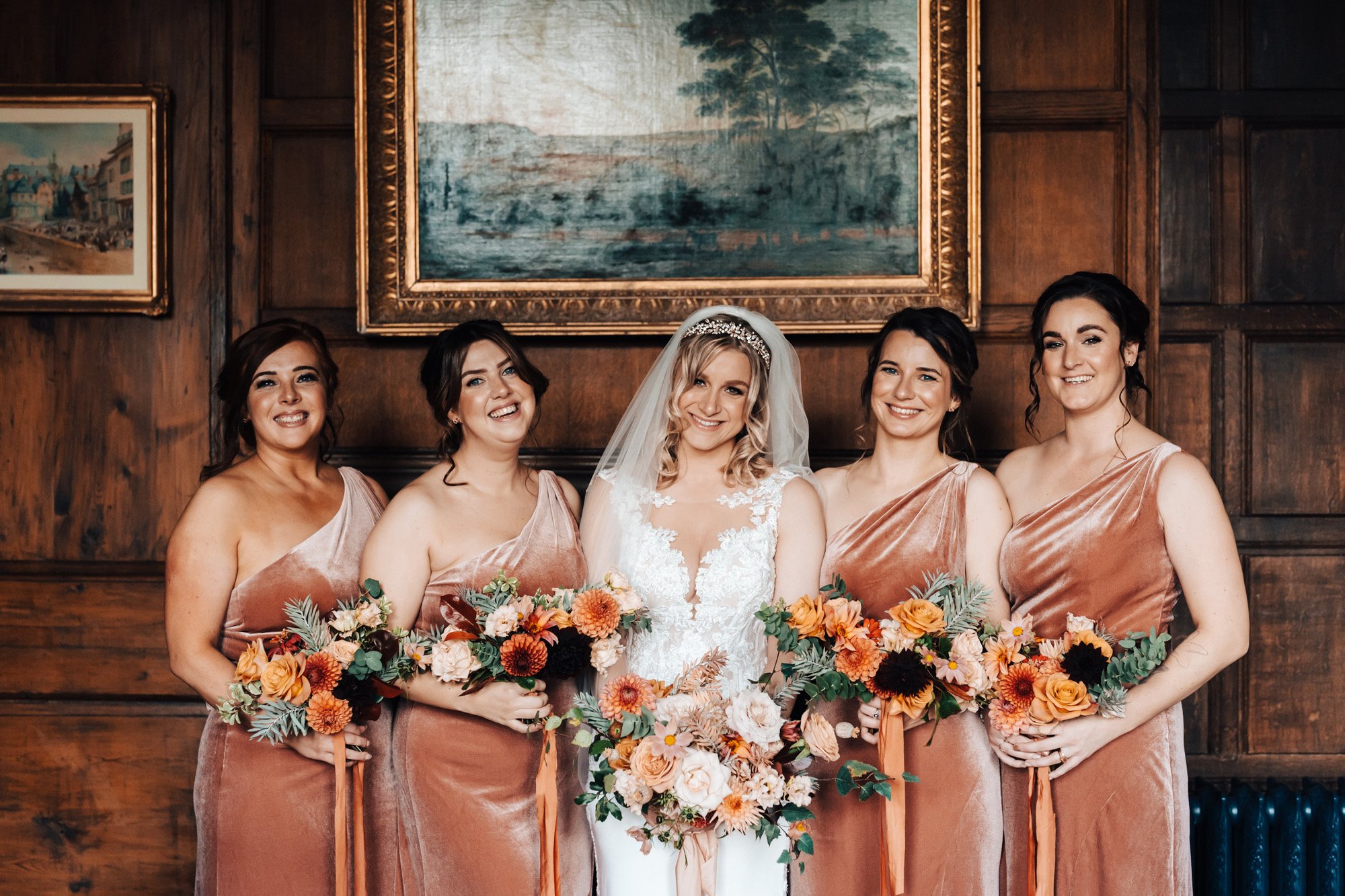 Bride and bridesmaids in pink velvet holding beautiful autumnal bouquets in a wood paneled stately home bedroom 