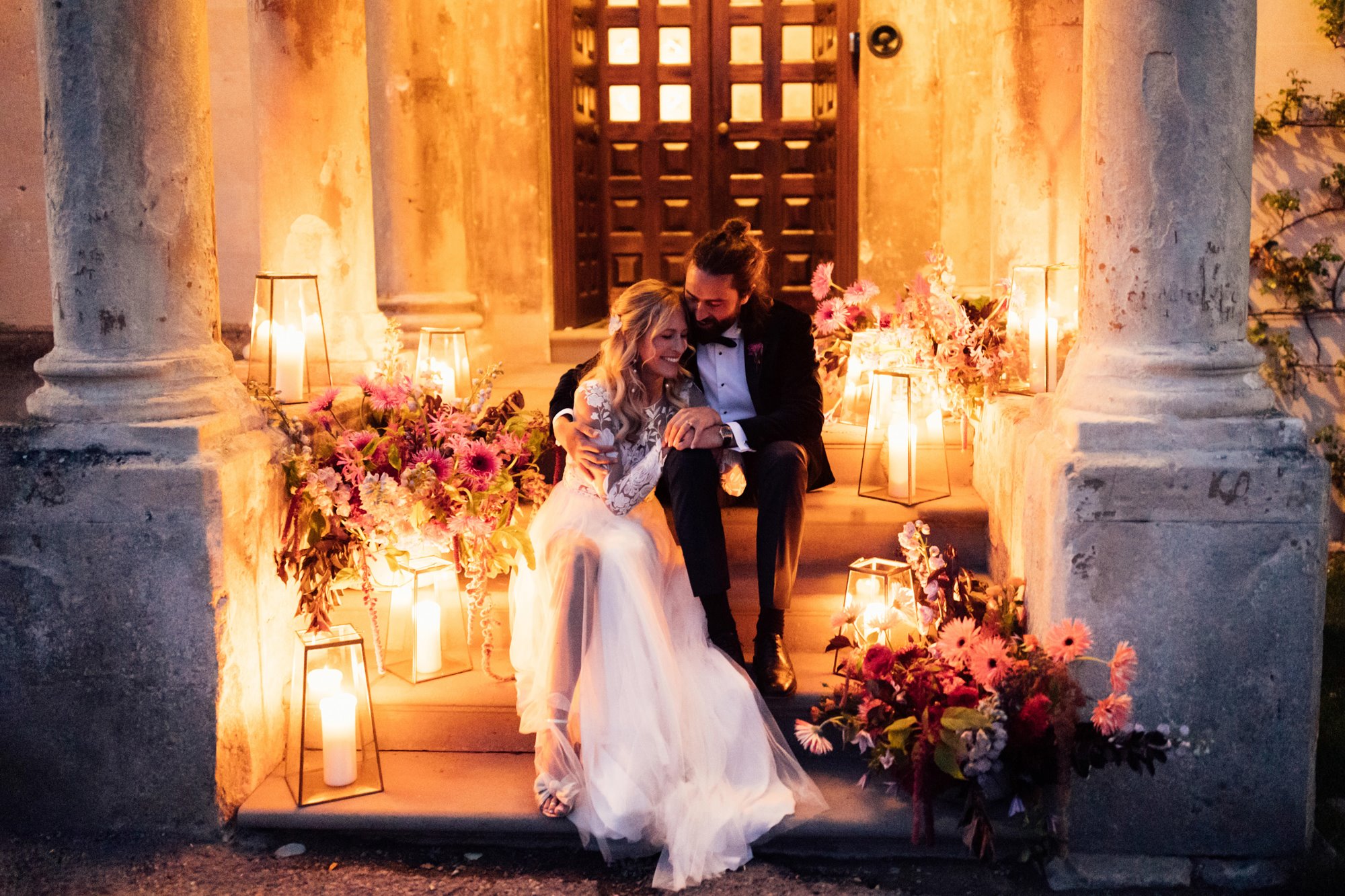 Bride and groom embrace on the steps of a stately home in Gloucestershire, surrounded by glowing candle light and stunning flower arrangements 