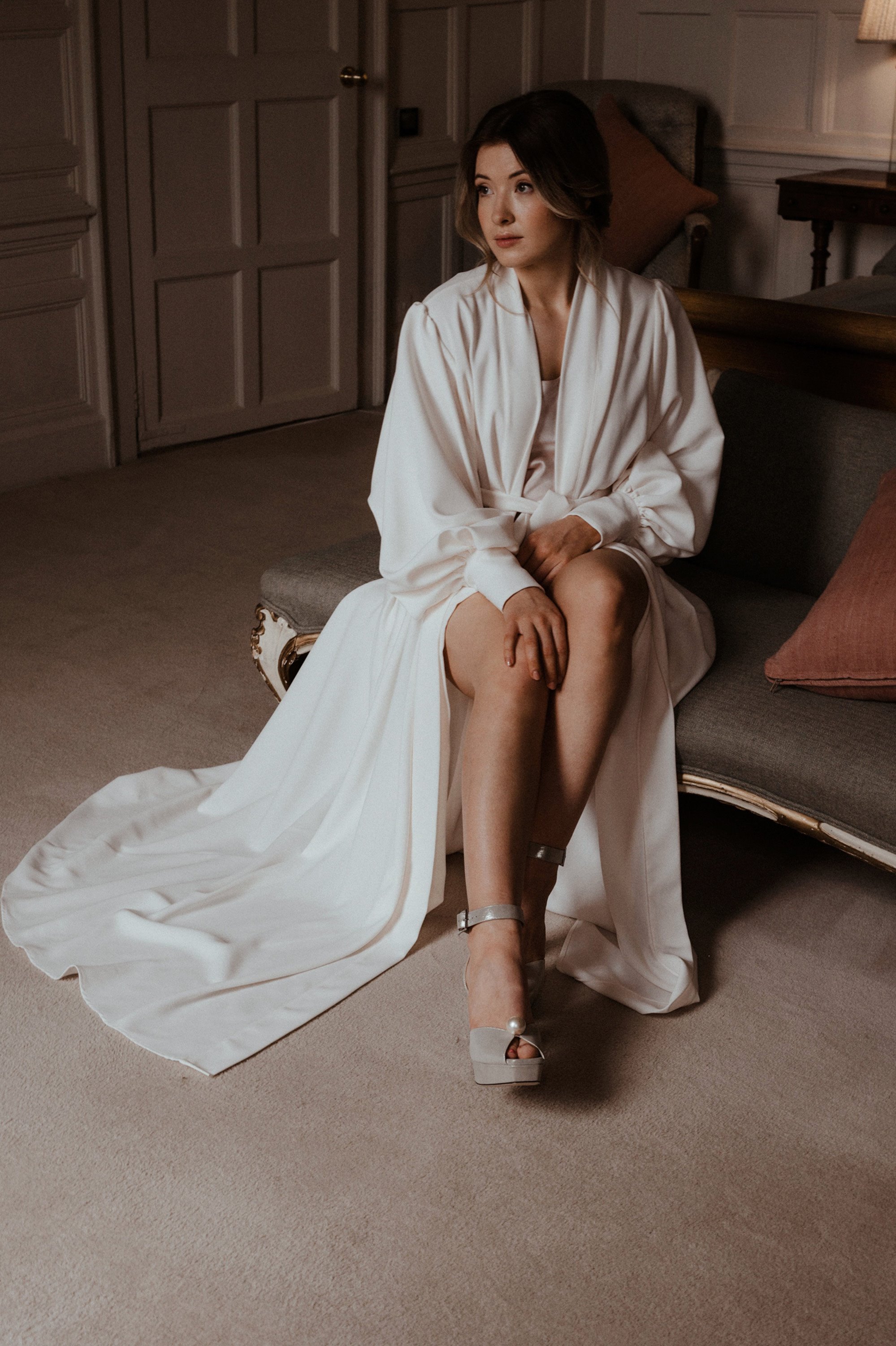 Bride in long white robe and puff sleeves wearing jimmy choo chunky wedges with strap and single pearl on toe sitting on chaise lounge in stately home bedroom