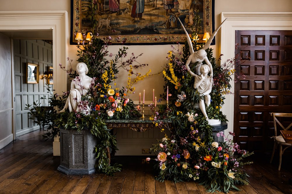Wild florals and greenery with candles decorate the wedding alter flanked by beautiful stone angel statues with wings in wedding ceremony hall at stately home elmore court in gloucester