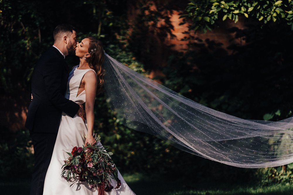 glamorous bride wearing cathedral length pearl studded veil and groom kiss in gardens at moody autumn wedding 