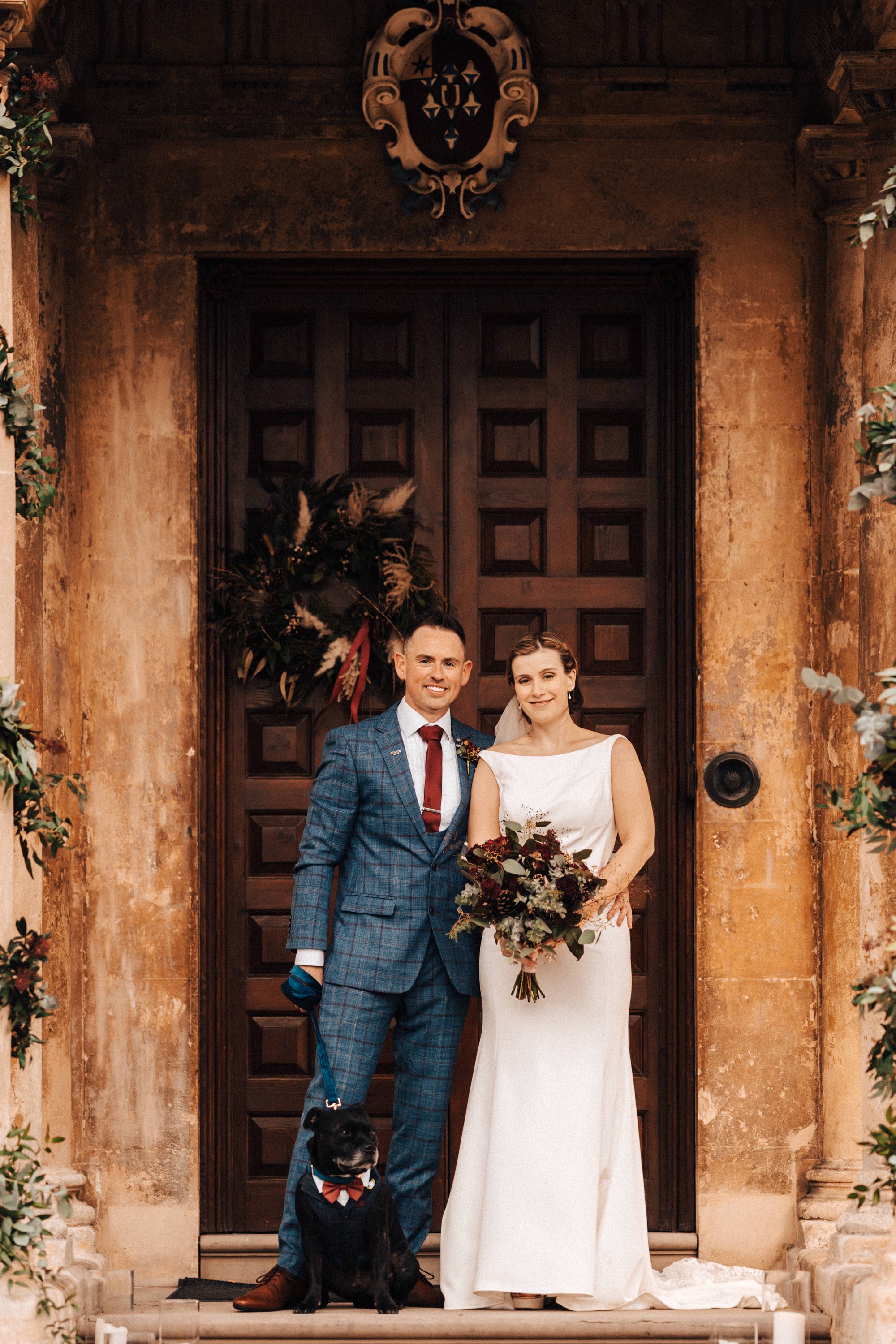 Bride and groom with dog outside their winter wedding venue with christmas wreath on the door