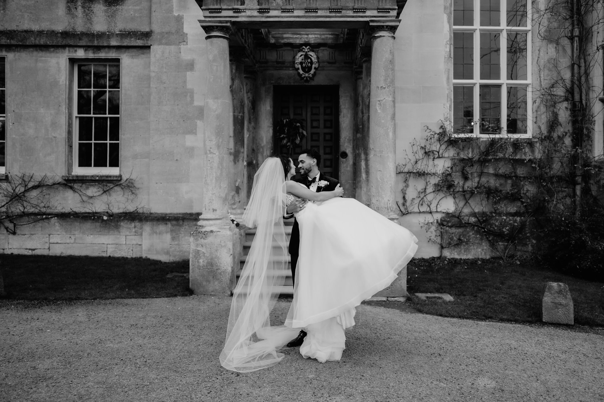black and shite shot of a Groom carrying his new wife outside a stately home in Gloucestershire