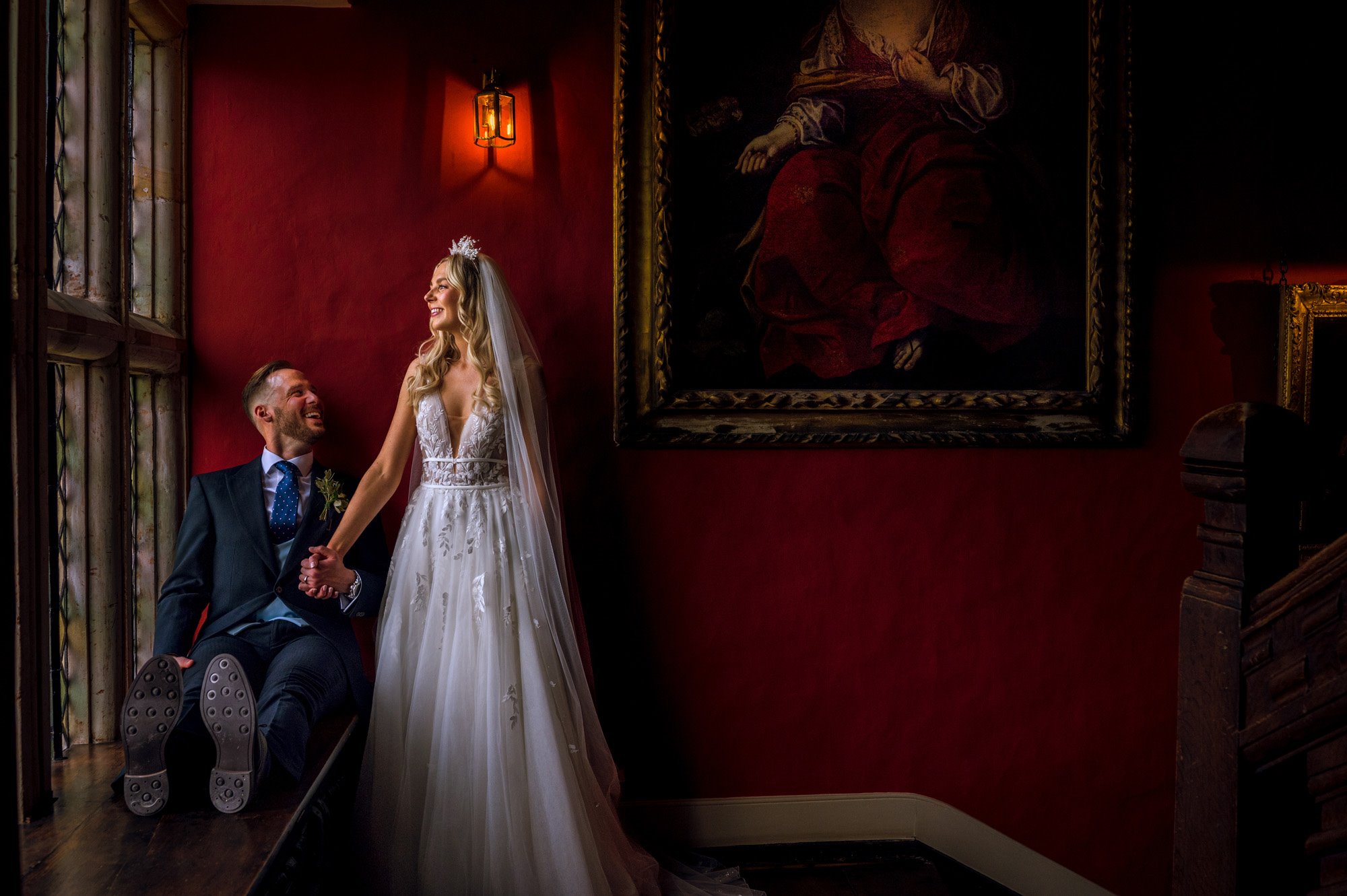 Bride and groom against striking red hallway of historic house wedding venue Elmore Court. Groom sits in huge windowsill and bride looks out in beautiful boho tulle dress and pearl crown