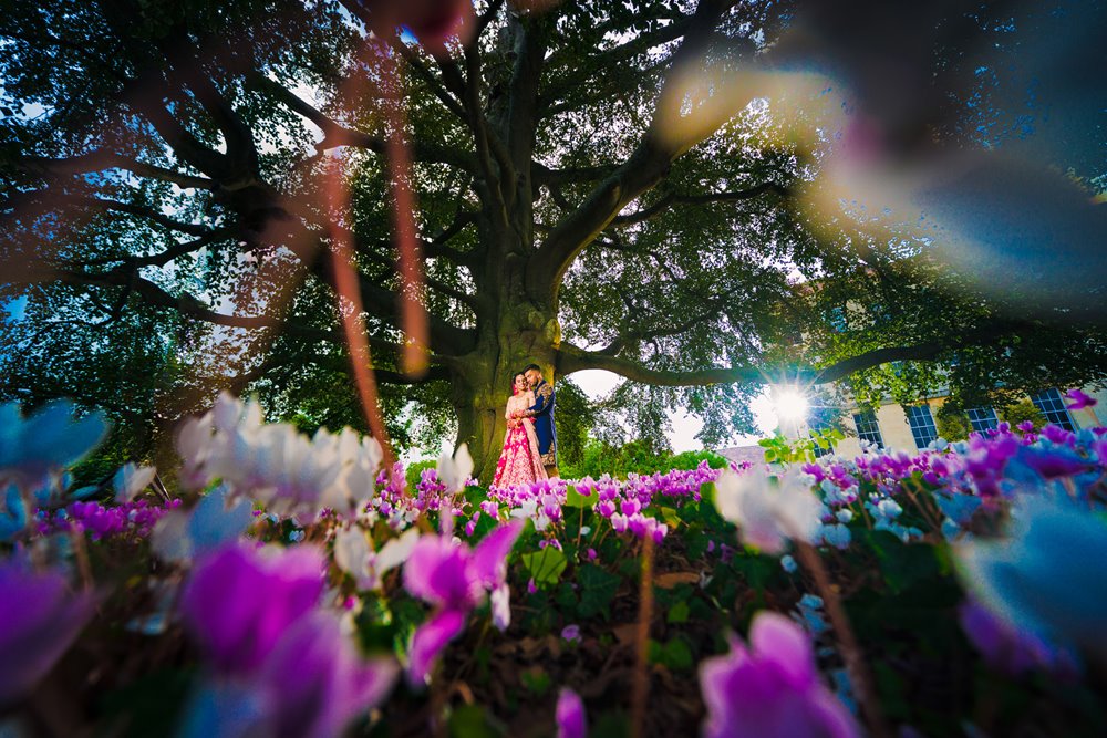 Indian bride and groom stand under old beech tree surrounded by flowers at elmore court