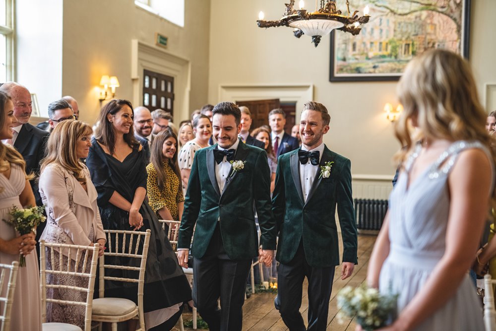 Two grooms nervously walking up the aisle together holding hands ready to become husband and husband at their beautiful gay wedding ceremony in stately home elmore court